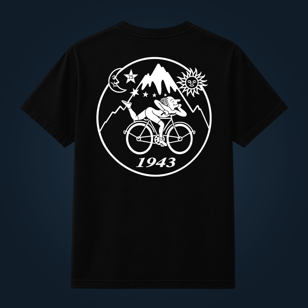 Circular graphic on a black tee featuring a cyclist riding uphill within mountains, sun and moon imagery, and the year 1943 in script below.